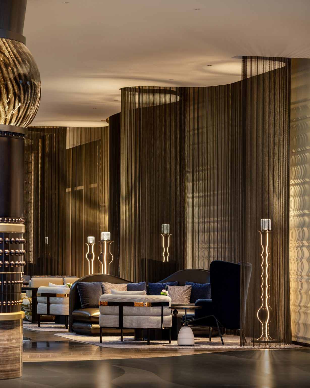 Luxurious hotel lounge with dim lighting, featuring elegant blue and beige sofas, sleek tall lamps, and floor-to-ceiling sheer curtains, creating a cozy ambiance.