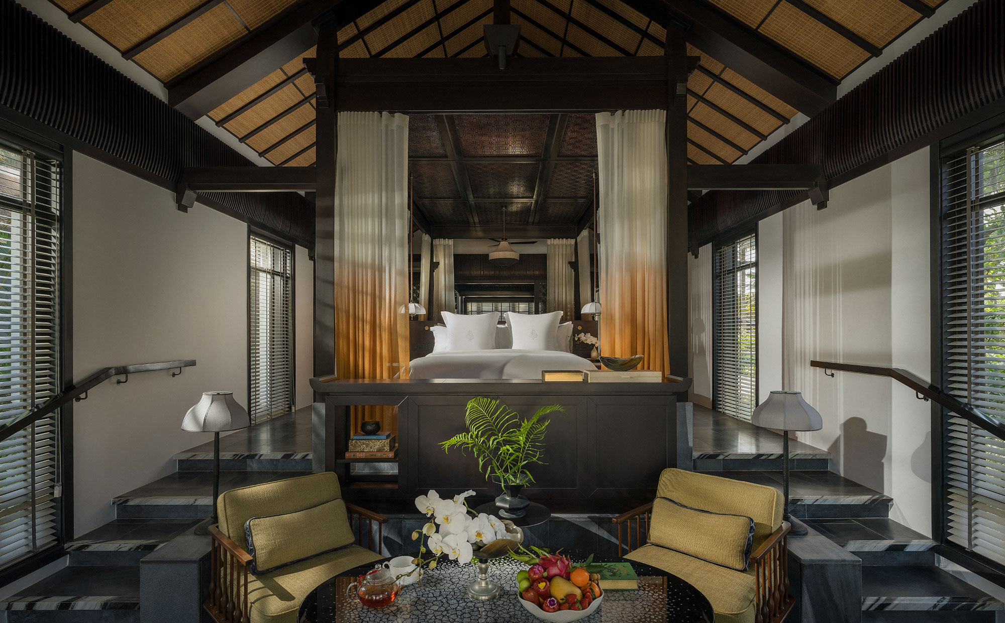 Grand resort villa master bedroom under a high vaulted wooden ceiling, featuring an elevated platform bed with sheer ombre drapes, flanked by symmetrical steps leading down to a lower seating area.
