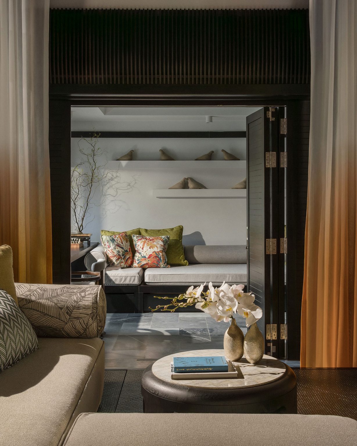 View through an open doorway of a luxury resort villa, showing a lounge area with floral throw pillows, a marble-top coffee table with orchids, and elegant gold-to-white gradient curtains.