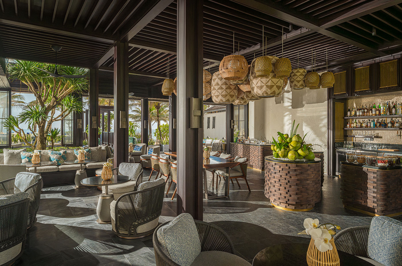 Sunlit luxury resort bar interior featuring woven wicker pendant lights, dark wooden pillars, comfortable lounge seating, and a fresh green coconut display on a round wooden counter.
