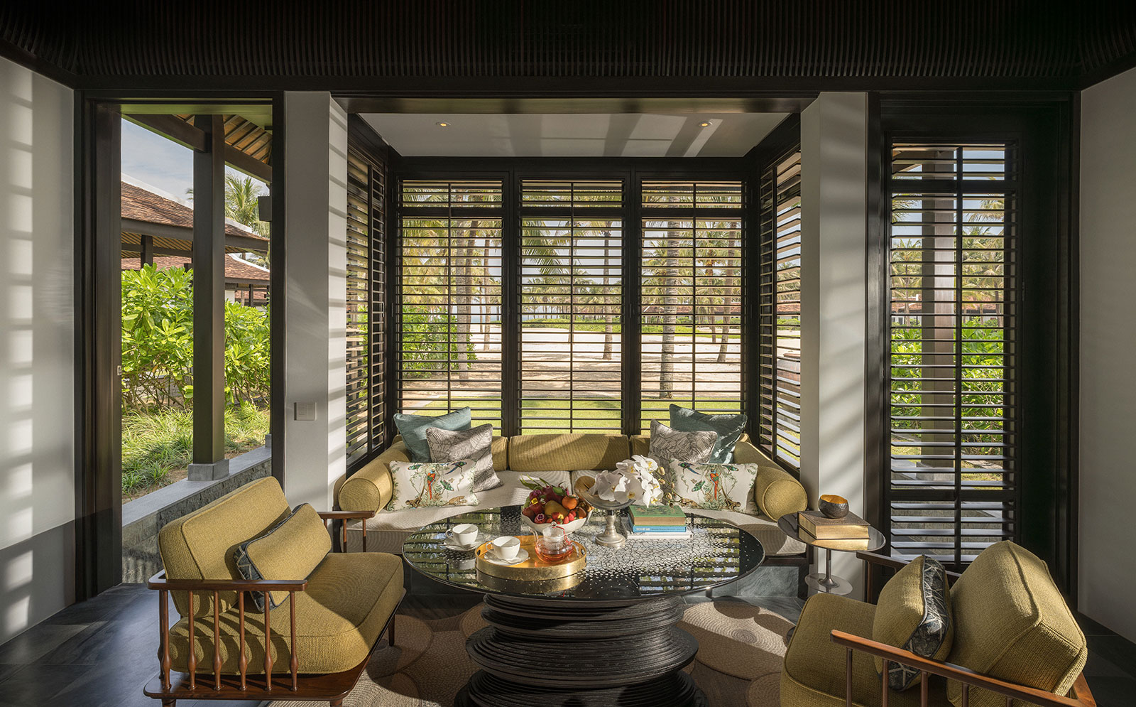 Sun-drenched villa sitting room with dark wood window shutters revealing tropical palm trees outside, furnished with green armchairs and a built-in daybed surrounding a round black coffee table.
