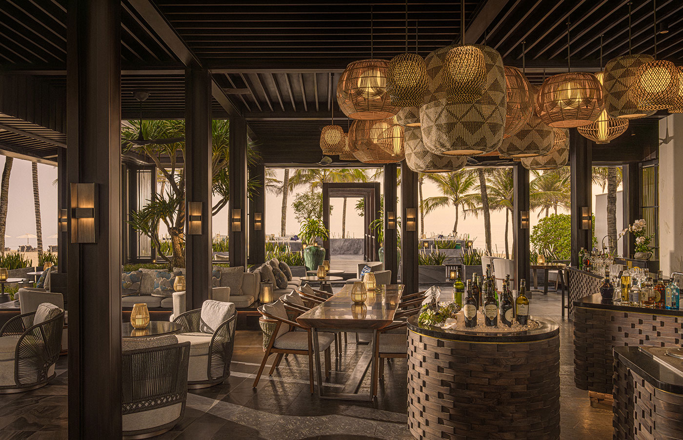 Warmly lit evening ambiance at a resort bar featuring large woven basket pendant lamps, dark wood architectural columns, and an open layout looking out toward a palm tree-lined beach.
