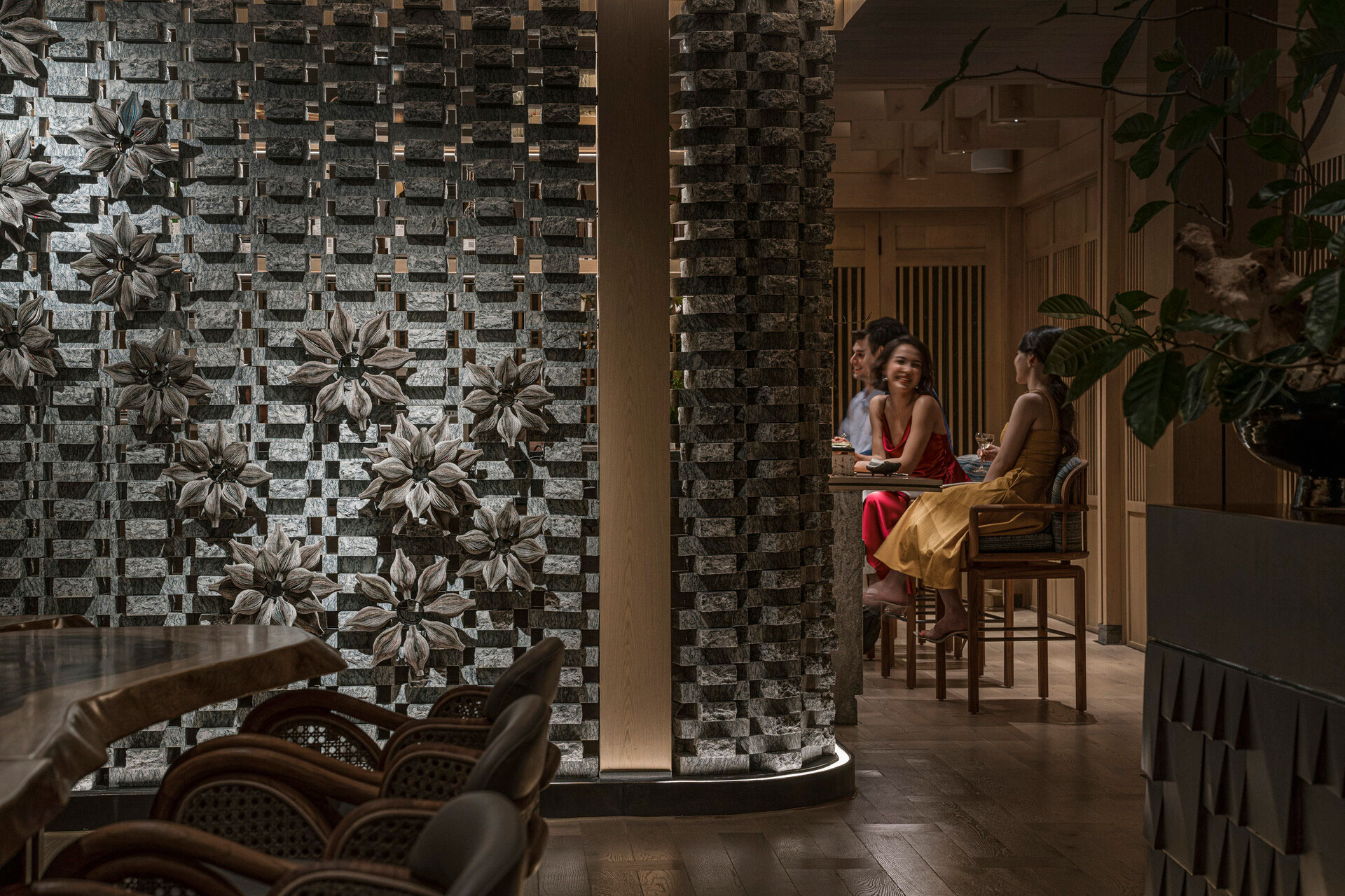 Guests enjoying a drink in a dimly lit, upscale Japanese restaurant featuring a decorative brick textured accent wall adorned with metallic flower sculptures.