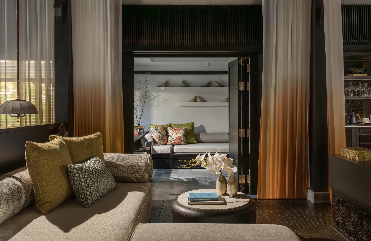 Elegantly styled villa living area with a plush sofa, patterned throw pillows, and ombre gradient curtains opening up to a separate lounge room in the background.