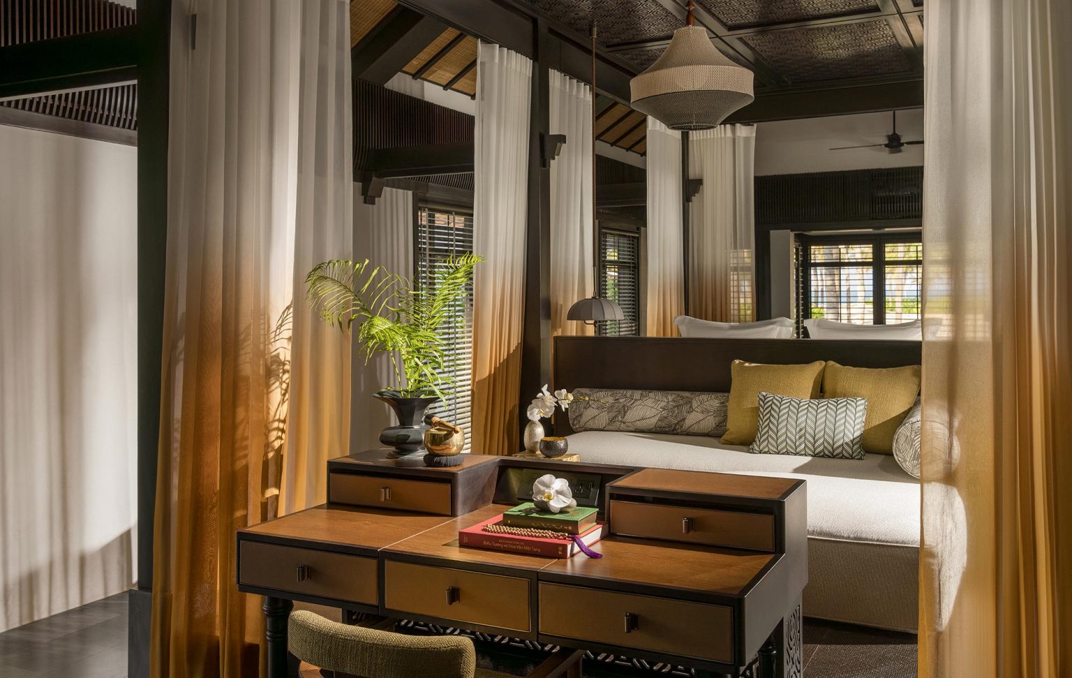 High-end villa interior showcasing a classic wooden writing desk with drawers, framed by translucent gold-to-white gradient curtains and a cozy daybed in the background.