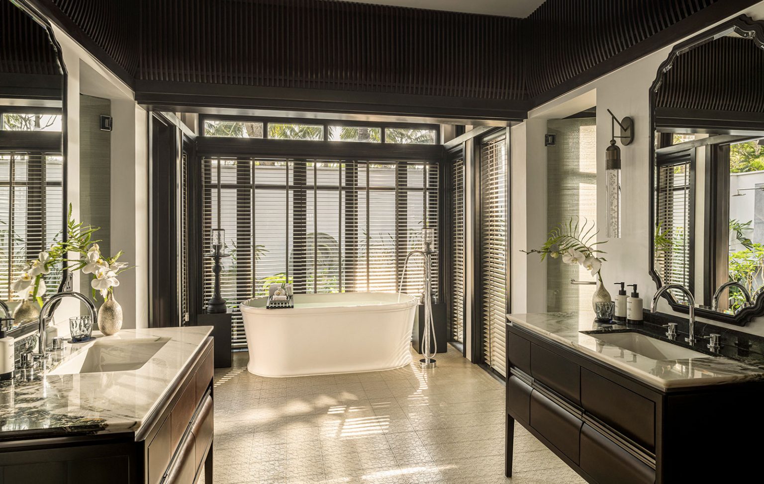 Spacious luxury bathroom featuring a freestanding white soaking tub by a large shuttered window, flanked by two dark wood vanities with marble countertops, mirrors, and elegant white orchid floral arrangements.