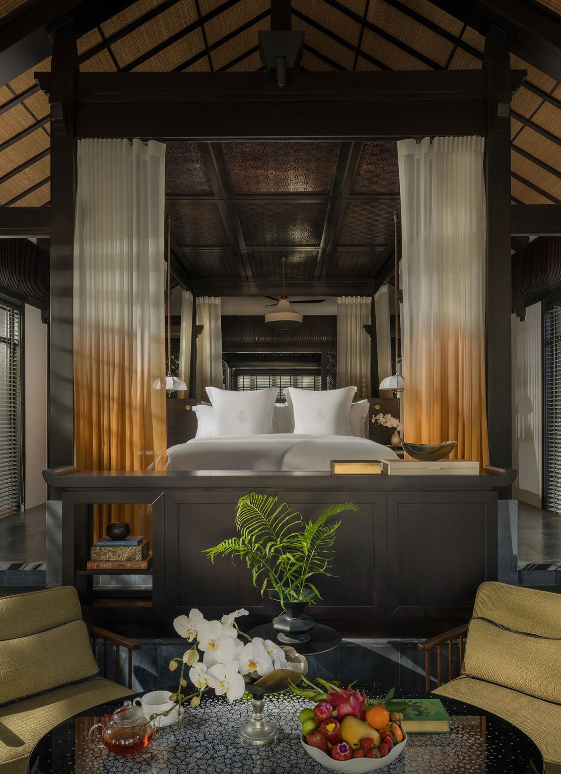 Grand resort villa master bedroom under a high vaulted wooden ceiling, featuring an elevated platform bed with sheer ombre drapes, flanked by symmetrical steps leading down to a lower seating area.