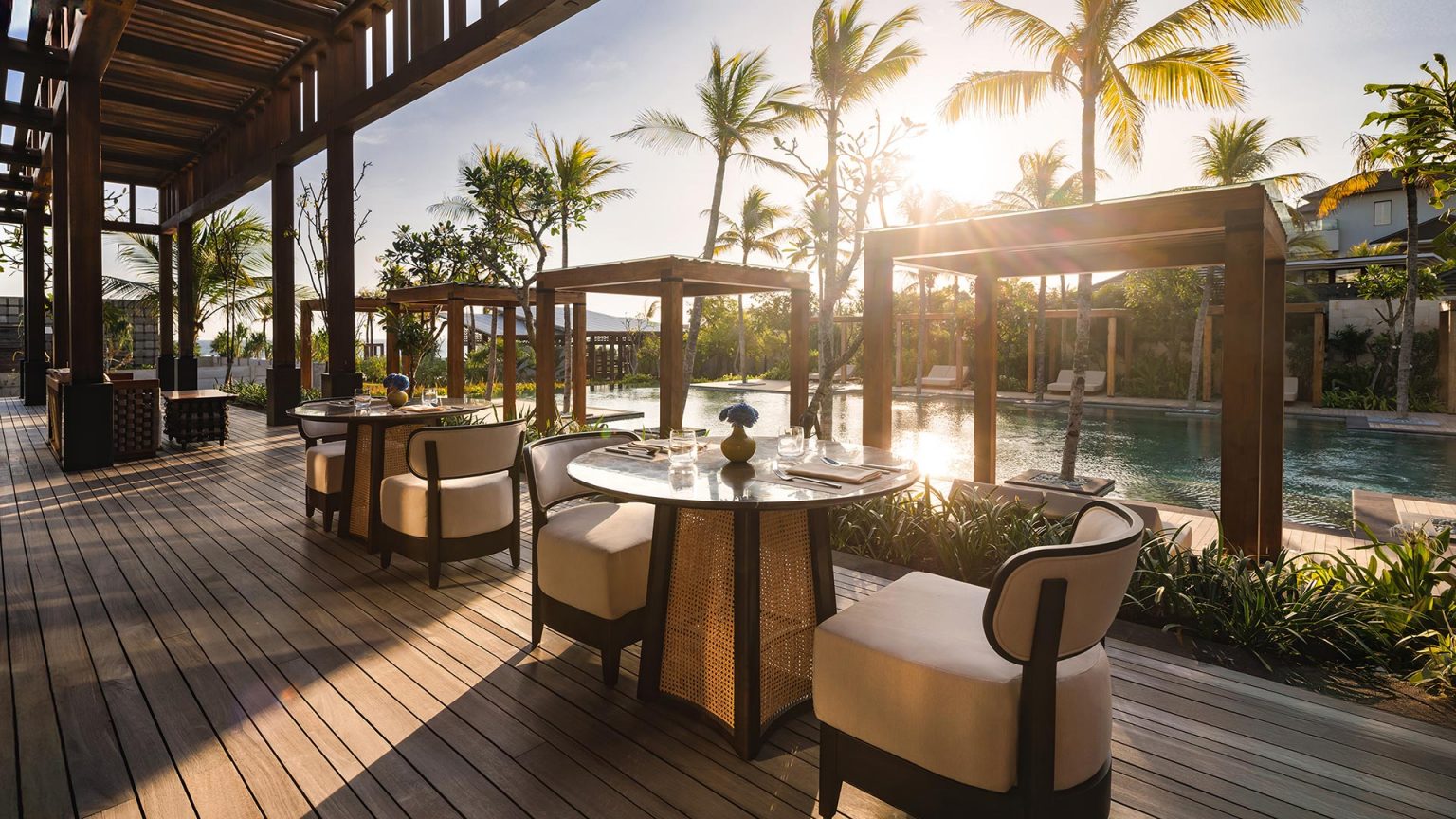 Outdoor dining area overlooking a sunlit pool, surrounded by palm trees. Wooden tables and cushioned chairs create a relaxing, tropical ambiance.